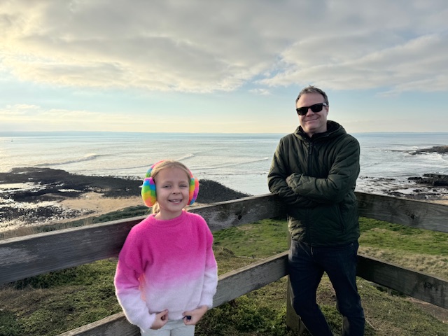 A photo of a man in a warm winter jacket. He's wearing sunglasses. Beside him is a young girl in a bright pink jumper. Behind them is the water of a beautiful bay.
